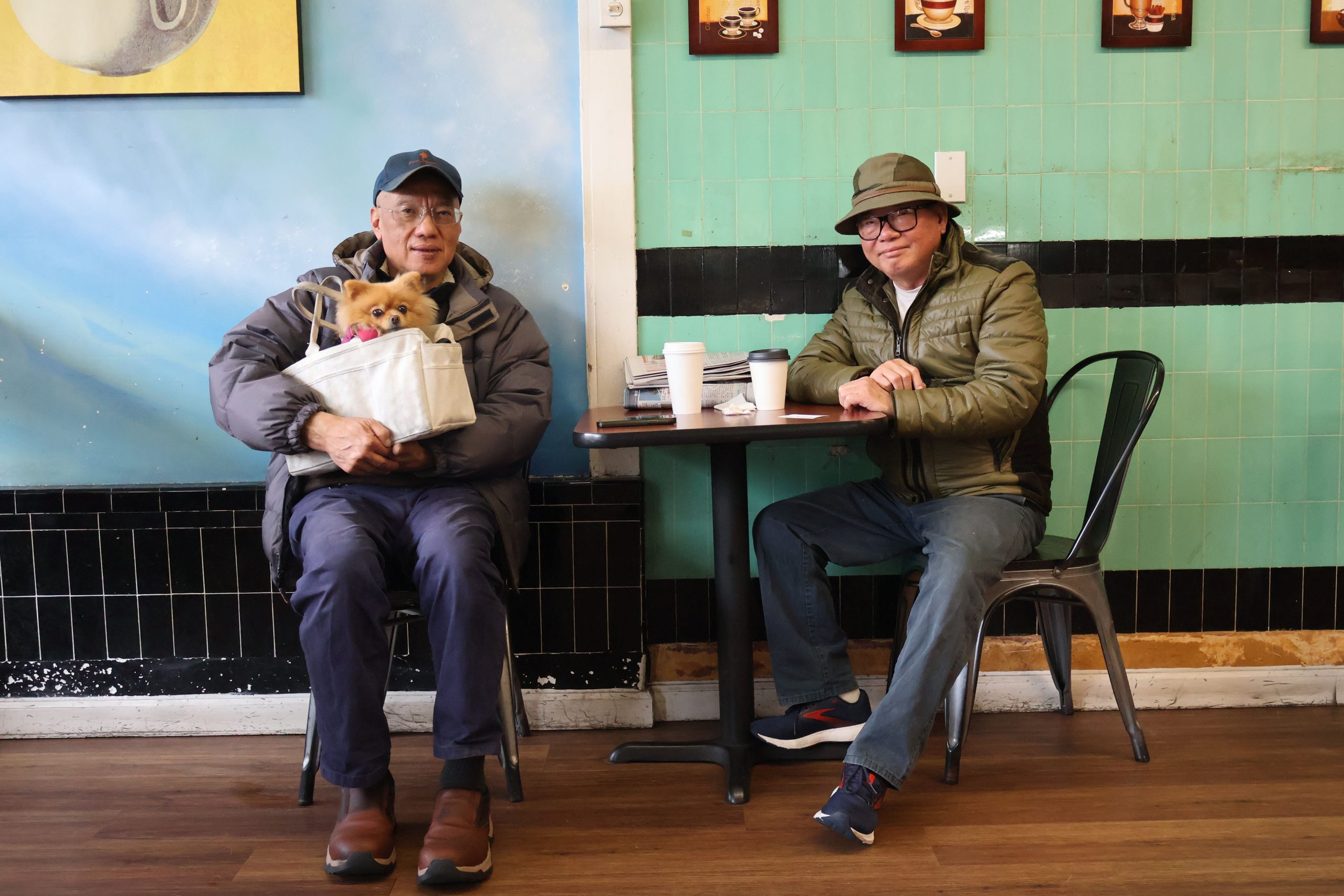 两名中国男子分别坐在咖啡馆的两张桌子旁，其中一人怀里抱着一只小狗。