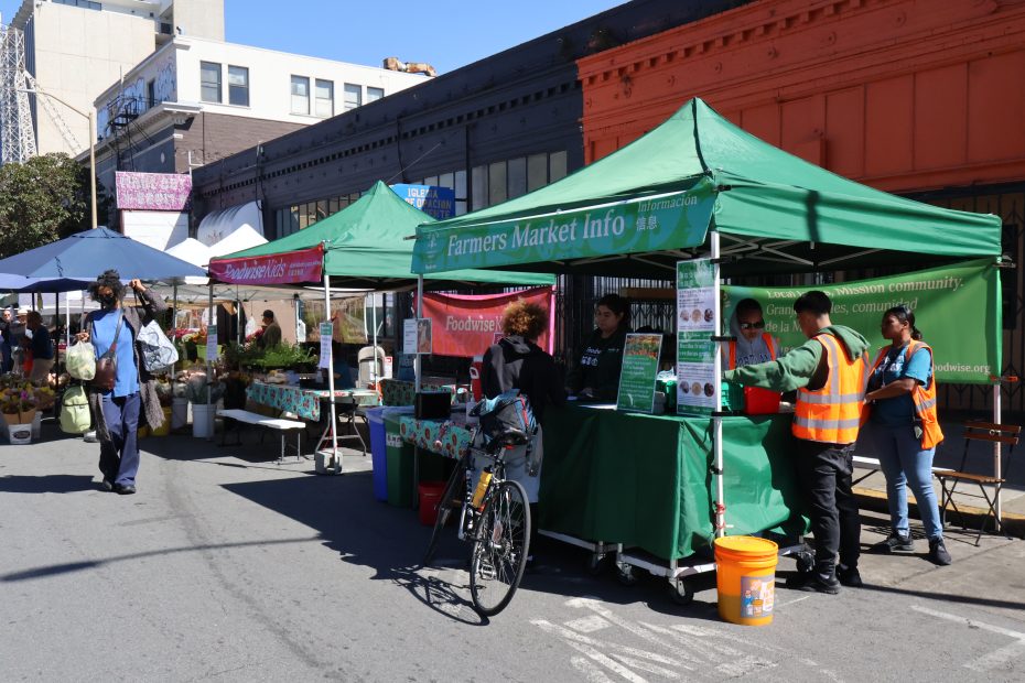 Outdoor farmers market match scene with people visiting various stalls under green tents, one of which is an information booth.