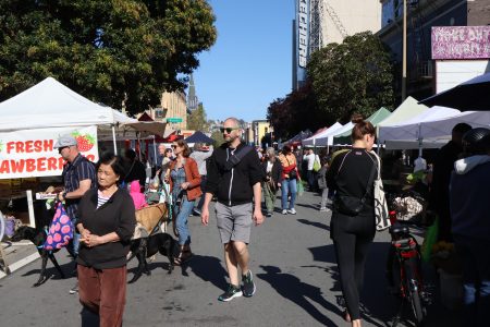 People walking and shopping at a busy outdoor market with various vendor tents displaying goods like fresh strawberries.