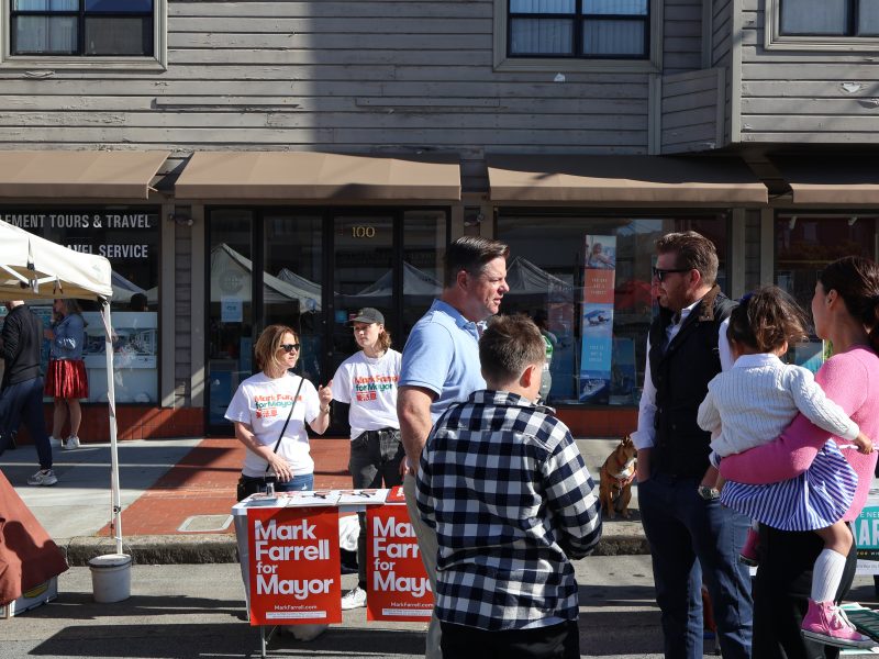 A man campaigning for mayor, "mark farrell," speaks with potential voters at a booth on a sunny street, accompanied by supporters and passersby.