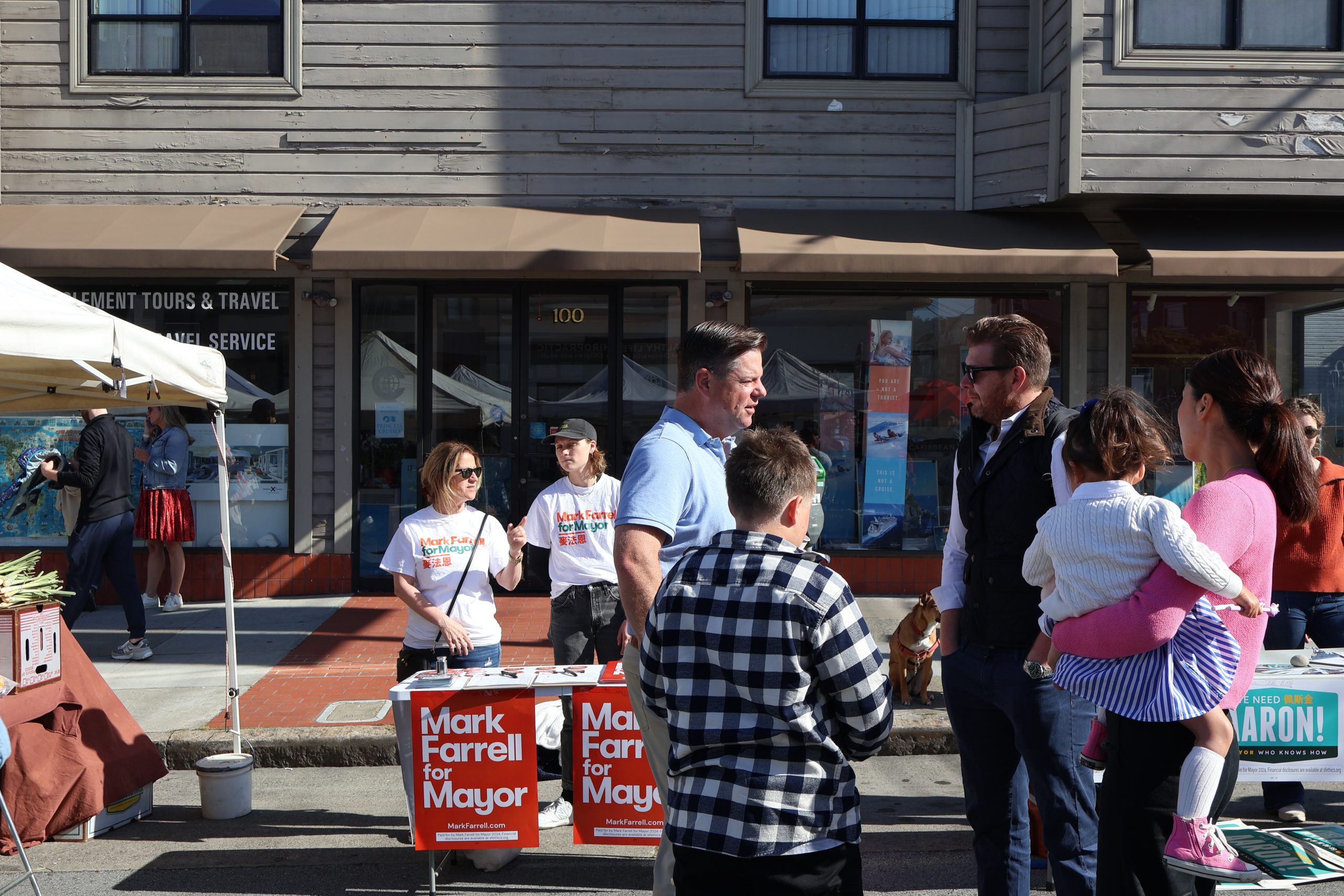A man campaigning for mayor, "mark farrell," speaks with potential voters at a booth on a sunny street, accompanied by supporters and passersby.