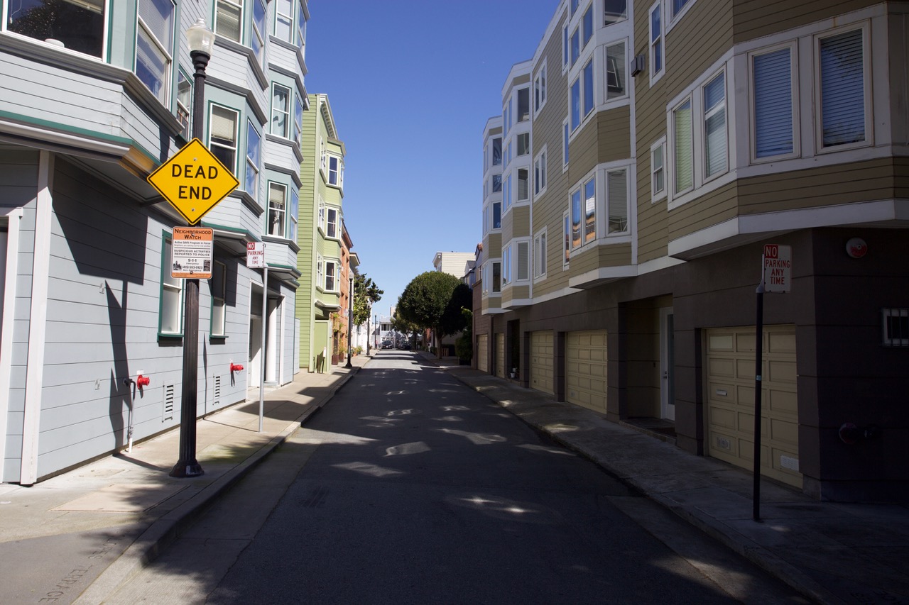 An empty street flanked by residential buildings with a 'dead end' sign visible under a clear sky.