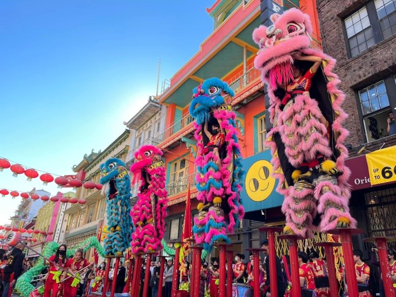 Colorful LionDanceME performance during a street festival with performers in traditional costumes holding up lion figures against a backdrop of buildings and red lanterns.