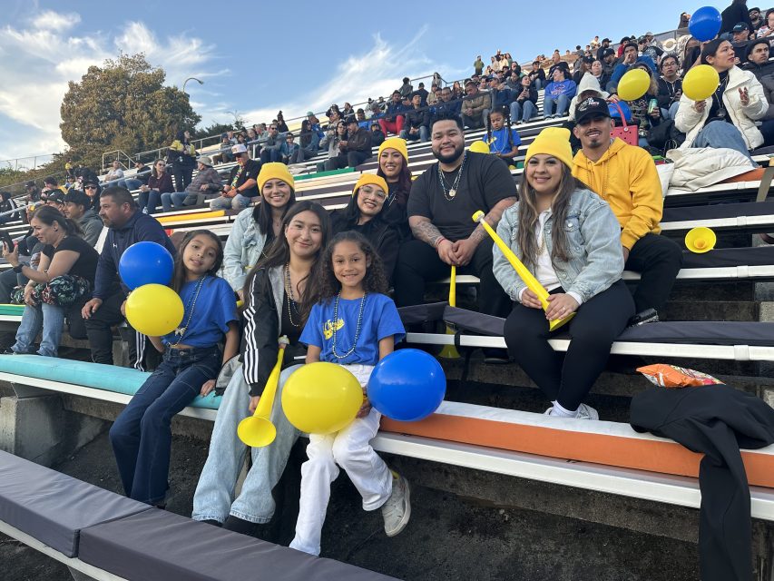 Group of people sitting on stadium bleachers, smiling and holding yellow balloons, wearing casual clothing and sports accessories.