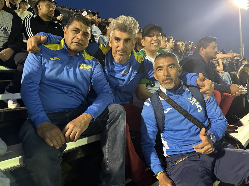 Four men in blue sports jackets sitting in El Farolito stadium seats at night, smiling and looking at the camera.
