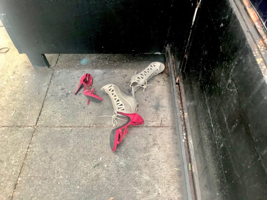 Two pairs of high-heeled shoes, one red and one white with cut-out patterns, abandoned on a sidewalk next to a black wall.