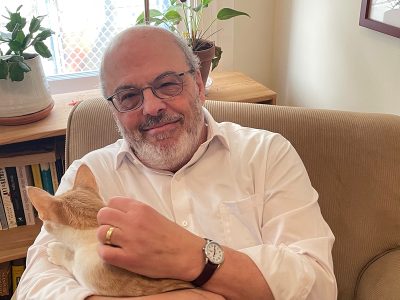 A smiling man with glasses and a white shirt, sitting indoors and holding an orange cat in his lap.