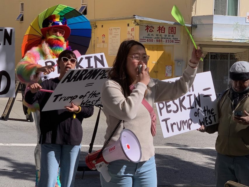 A group of protesters, one holding a rainbow umbrella, display signs. A woman speaks into a megaphone in the foreground. Signs read "Peskin = Trump of the West" and "AARON PESKIN = Trump of the West.