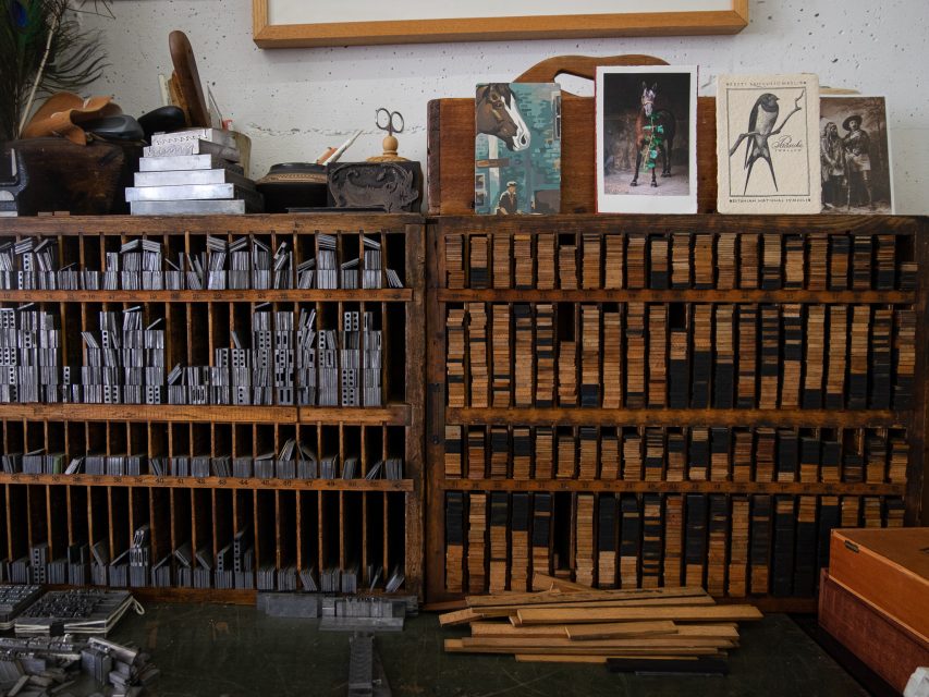 A vintage print studio featuring rows of wooden letterpress drawers filled with typeset blocks, alongside various printing tools and framed artworks on display.