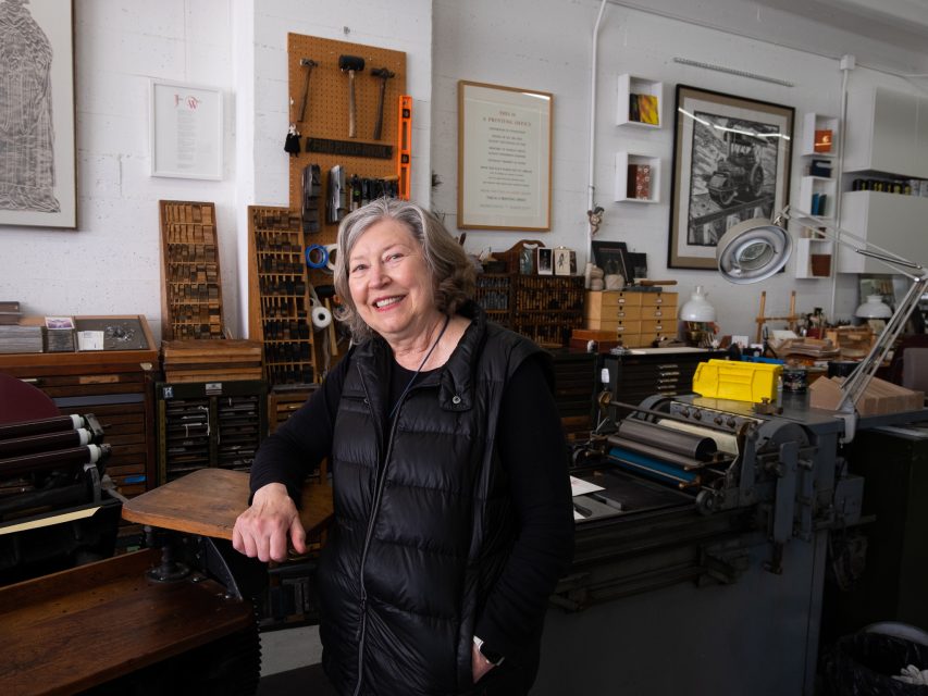 A smiling older woman standing in a print workshop surrounded by tools, printing presses, and typeset drawers.