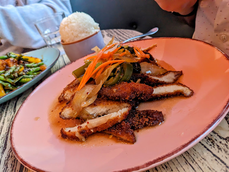 A person holding a plate of breaded pork cutlets topped with sautéed onions and julienne vegetables, accompanied by a scoop of rice, in a restaurant setting.