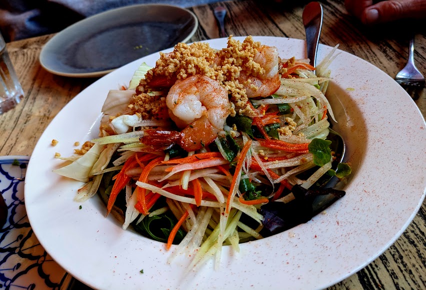 A plate of thai green papaya salad with shrimp, julienned vegetables, herbs, and crushed peanuts, presented on a wooden table.