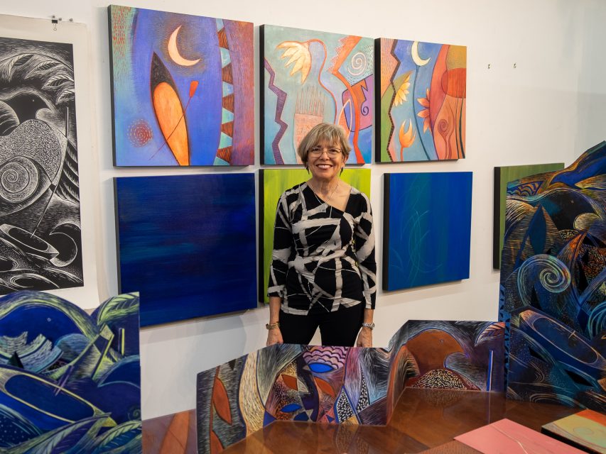 A smiling woman stands in front of vibrant, colorful abstract paintings displayed in an art gallery.
