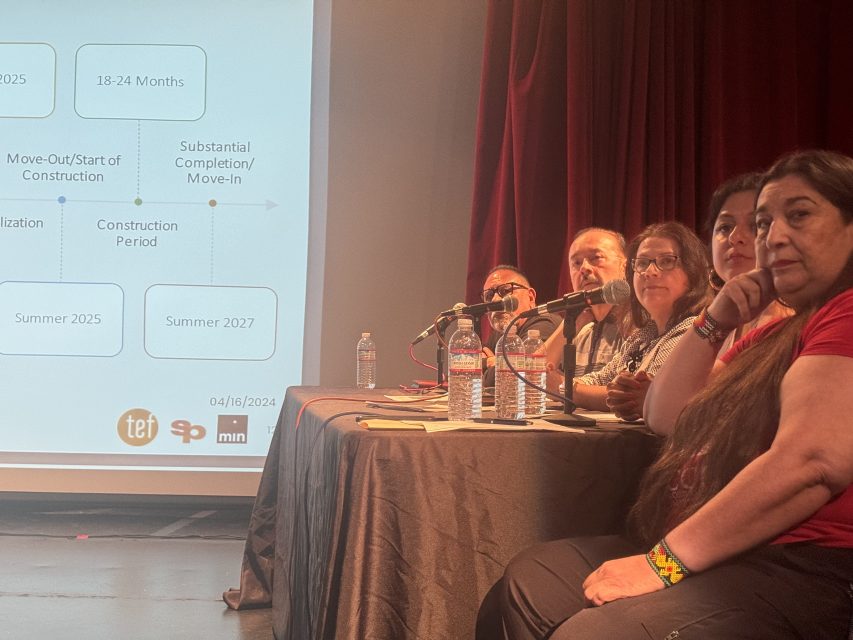 Panel of five people seated at a table with microphones during a public meeting with a presentation slide projected in the background.