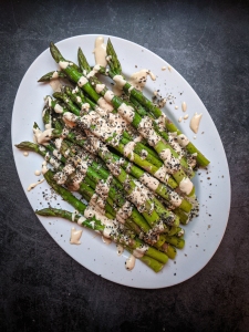 Oval plate of grilled asparagus garnished with sesame seeds, herbs, and drizzled with a creamy dressing, on a dark textured background.