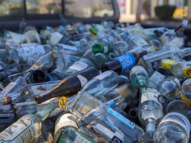 A pile of various discarded glass bottles in a recycling bin, focusing on their mixed colors and labels.