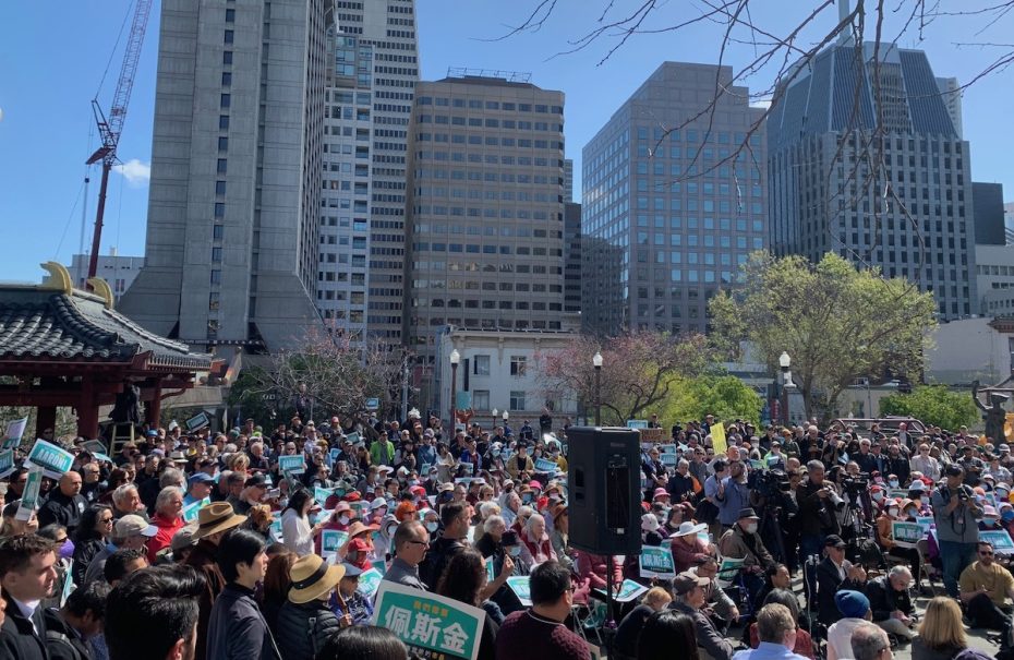 A large crowd gathers in the Peskin Square surrounded by tall buildings, holding various placards during a daytime event.