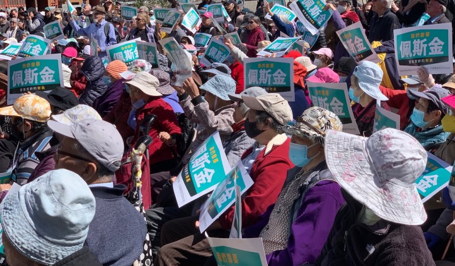 A large crowd of people, wearing hats and masks, hold up green and white signs while attending an outdoor event honoring Peskin.