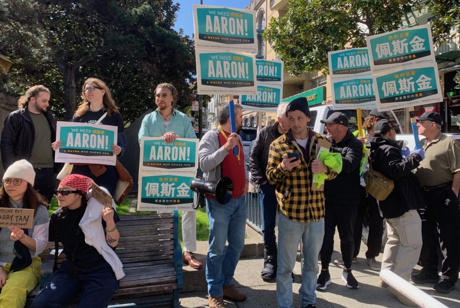 A group of people, including Peskin supporters, stands and sits, holding up signs that read "We need Aaron!" and other text in both English and Chinese on a sunny day outdoors.