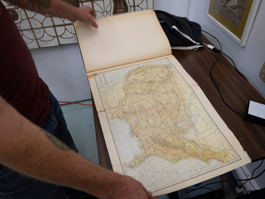A person turning the page of a large, vintage atlas book open to a colored map of a region, displayed on a table.