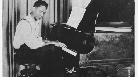 A man sits at a piano, looking at sheet music, in a vintage black-and-white photograph.