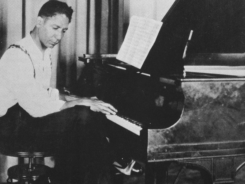 A man sits at a piano, looking at sheet music, in a vintage black-and-white photograph.