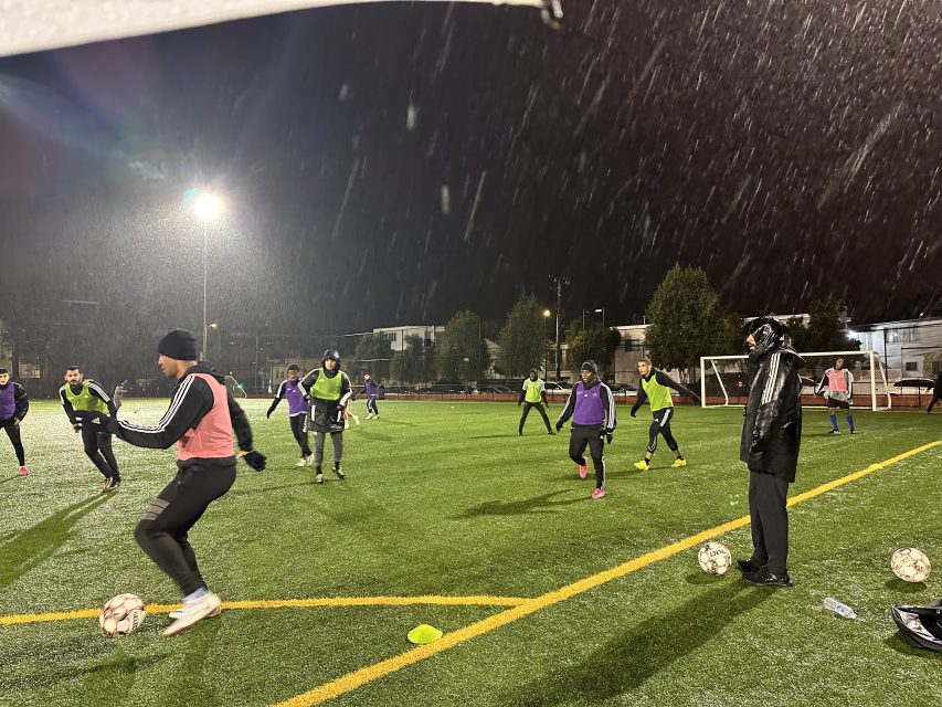 Adults playing soccer on a lit outdoor field during a nighttime snowfall, with an emphasis on motion and active participation.
