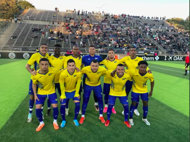 A youth soccer team dressed in yellow and blue uniforms posing for a group photo on a stadium field before a match.