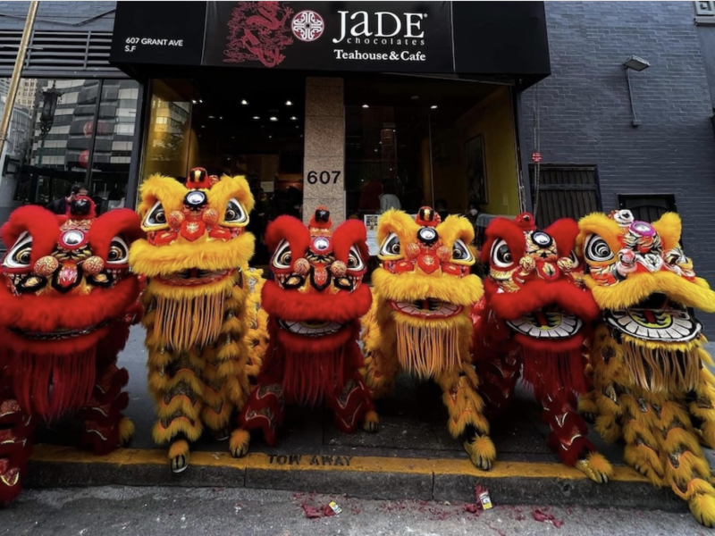 Performers in red and yellow lion dance costumes pose in front of the jade chocolates teahouse & cafe during a cultural celebration.