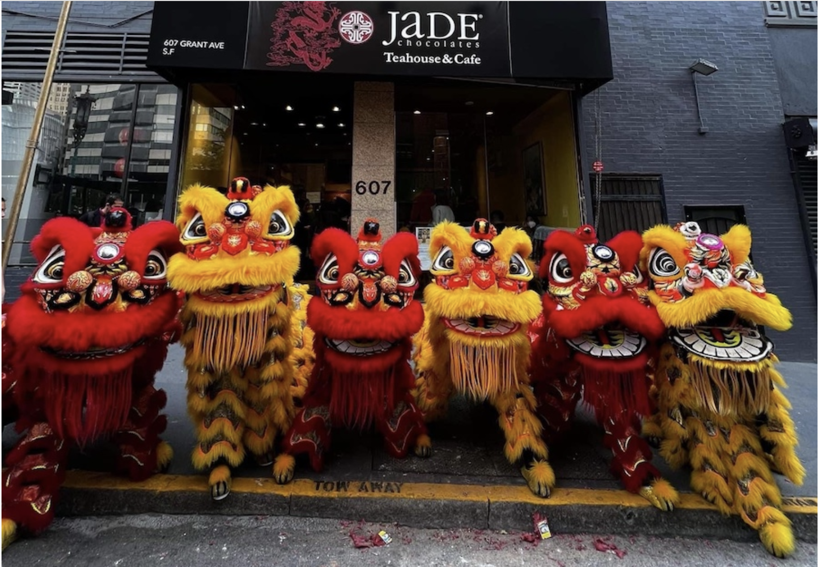 Performers in red and yellow lion dance costumes pose in front of the jade chocolates teahouse & cafe during a cultural celebration.