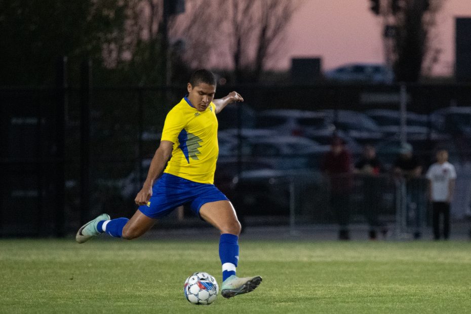 A soccer player in a yellow and blue uniform kicking a ball on a grassy field during an evening game.