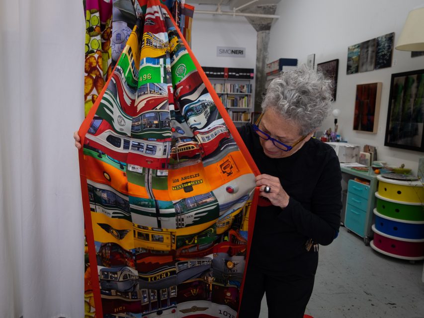 An older woman in a black outfit closely examines a colorful textile featuring a vibrant bus pattern in an art studio.