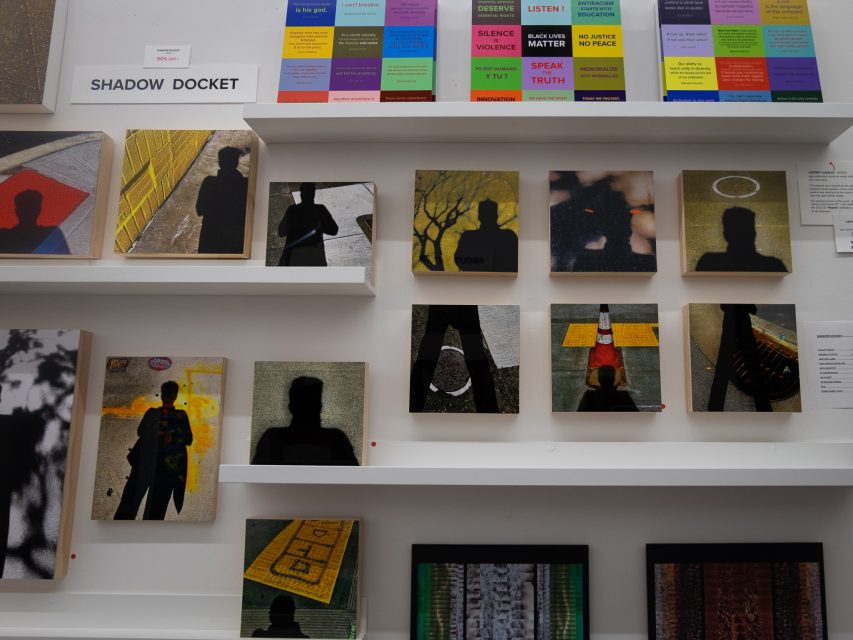 Art exhibition wall displaying various framed photographs and placards with text, arranged neatly on white shelves.