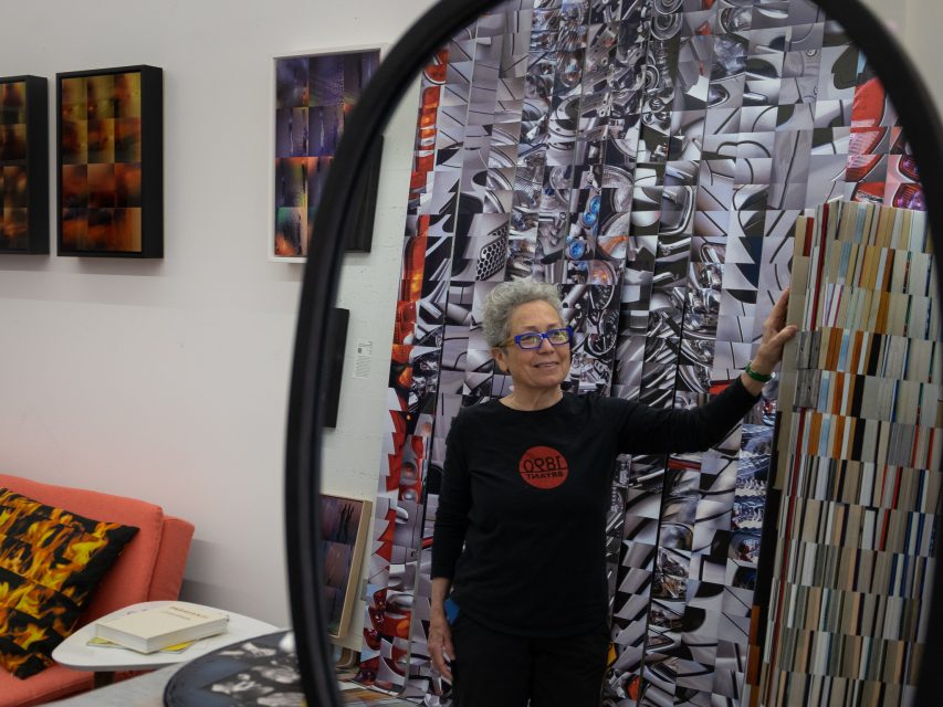 A woman with gray hair, wearing glasses and a black long-sleeve shirt, stands in an art studio, touching a colorful artwork.