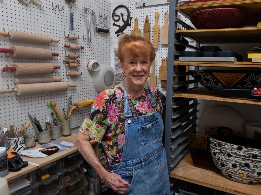 An elderly woman with red hair wearing a floral shirt and denim overalls stands in a well-organized craft room filled with tools and materials.
