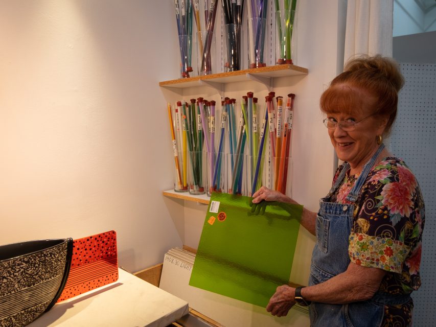 Elderly woman in an apron smiling while holding a green painting in an art studio surrounded by brushes and art supplies.
