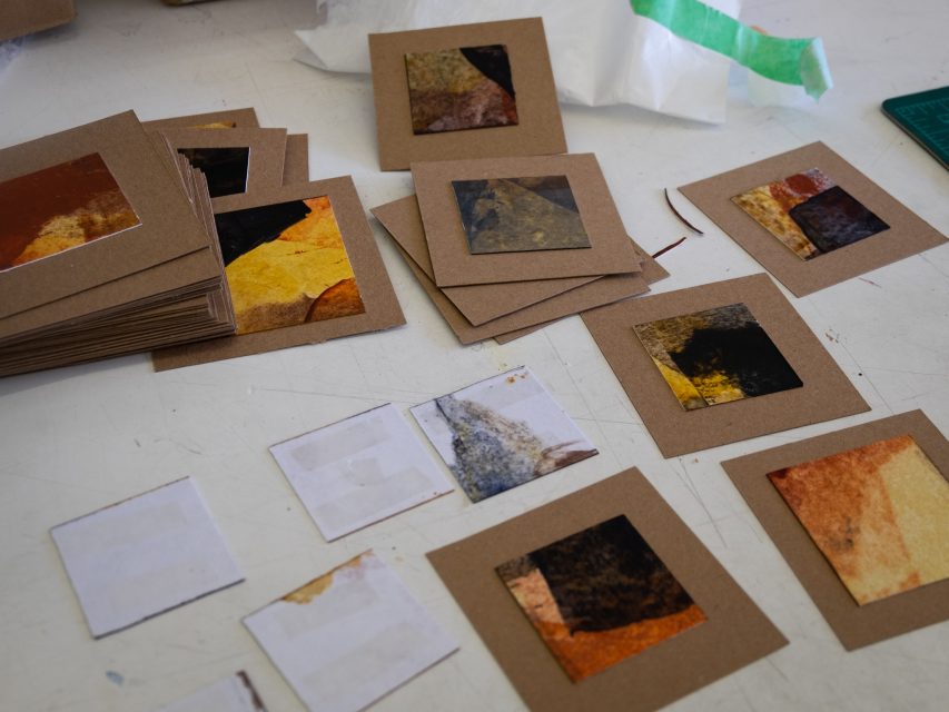 A collection of handmade cards with abstract, colorful designs spread out on a table in an art workshop setting.