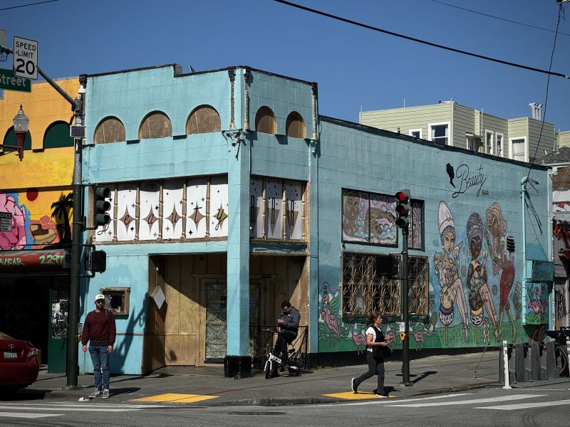 A teal building with boarded-up windows and colorful street art on its walls located at a sunny street corner, with pedestrians and traffic lights visible.
