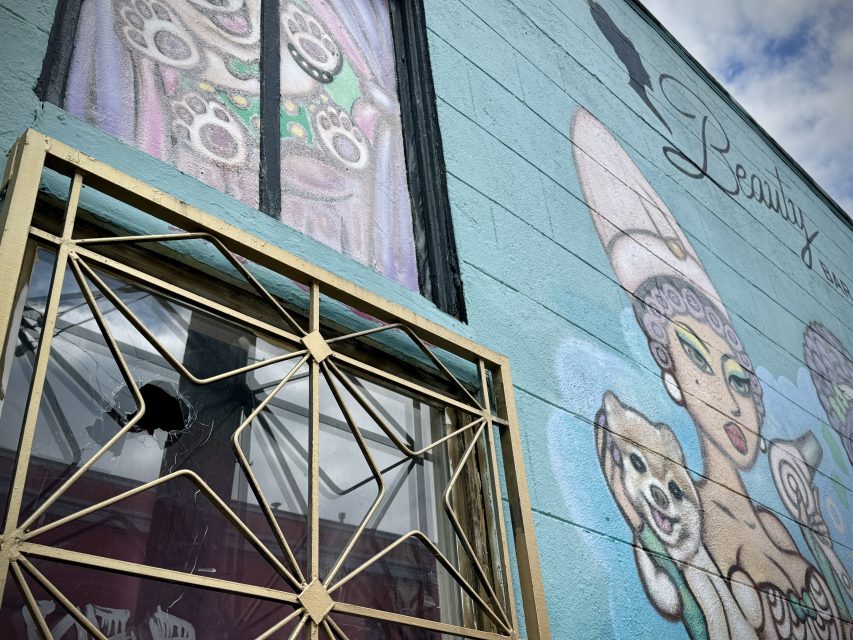 Mural of a stylized female figure on a building's wall with a broken window protected by a decorative metal grille.