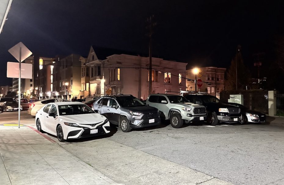 Nighttime street parking scene featuring several cars, including sedans and suvs, illuminated by streetlights in an urban area.