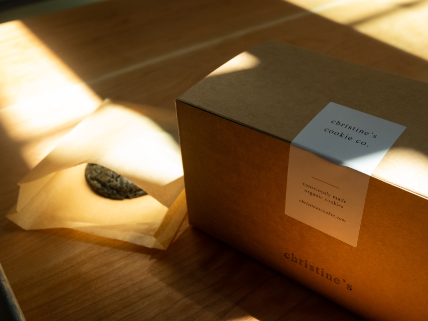 A cardboard cookie box labeled "christine's cookie co." in sunlit room, with a single cookie beside it on brown paper.