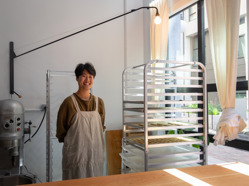 A smiling person in an apron stands next to a tray rack in a bakery kitchen, with a mixer and curtain-draped windows in the background.