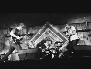 Black and white photo of a band performing on stage with a guitarist on the left, drummer in the center, and another guitarist on the right.