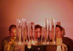 Three men looking through transparent, vertical rods on a table, which distort their faces, in a room with warm lighting.