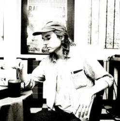 Black and white image of a young man in a newsboy cap sitting at a cafe table, looking to the side, with a drink and window in the background.