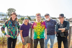 Five men posing outdoors, each with a unique style, standing by a waterside railing with greenery and buildings in the background.