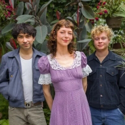 Three young adults smiling in a garden, with one woman in a purple dress flanked by two men in denim jackets.