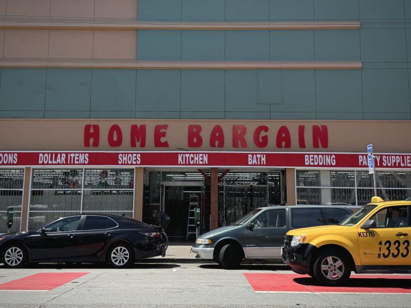 Exterior of the "home bargain" store with parked cars and a taxi, showcasing various product signs like shoes and bedding in the storefront windows.