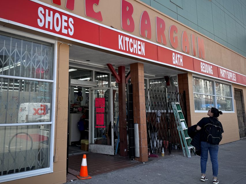 A person stands outside a bargain store featuring signs for shoes, kitchen, bath, and bedding products.