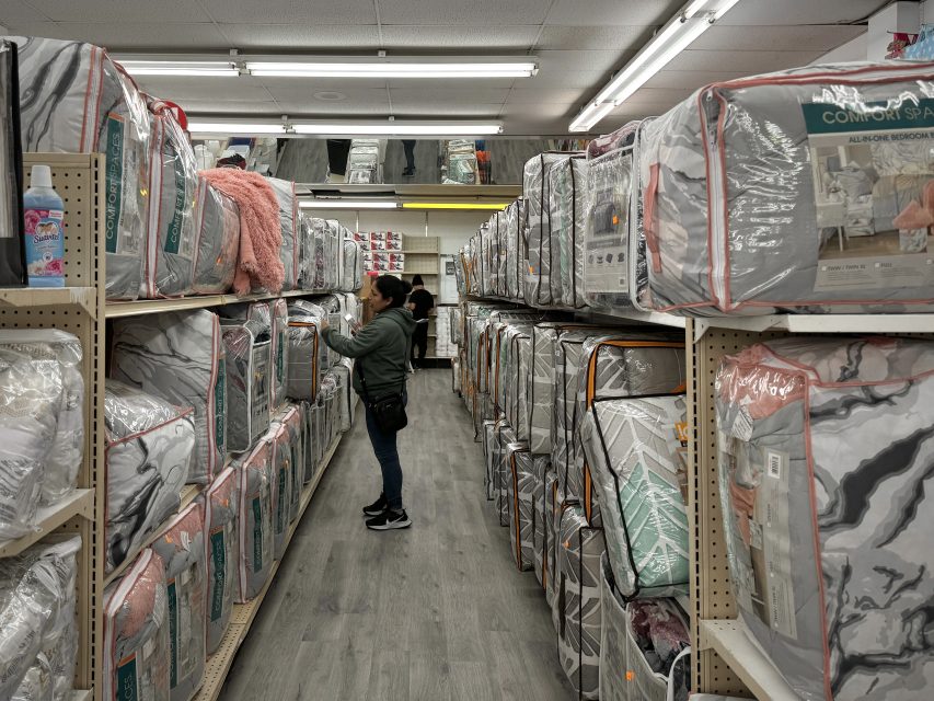 A woman browsing bedding items in a store aisle filled with shelves stocked with packaged comforters and bedspreads.